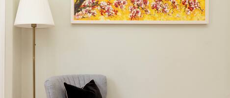 A room with a grey armchair, a black cushion, a floor lamp, and a framed painting of flowers on the wall.
