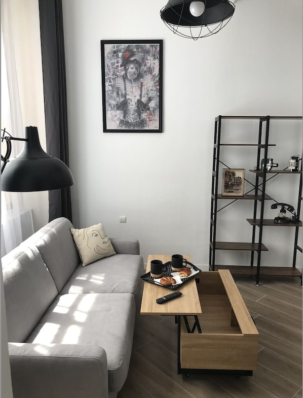 A modern living room with a grey sofa, a wooden coffee table with a tray of pastries, a black floor lamp, a framed abstract painting, and a metal shelving unit with a vintage camera.