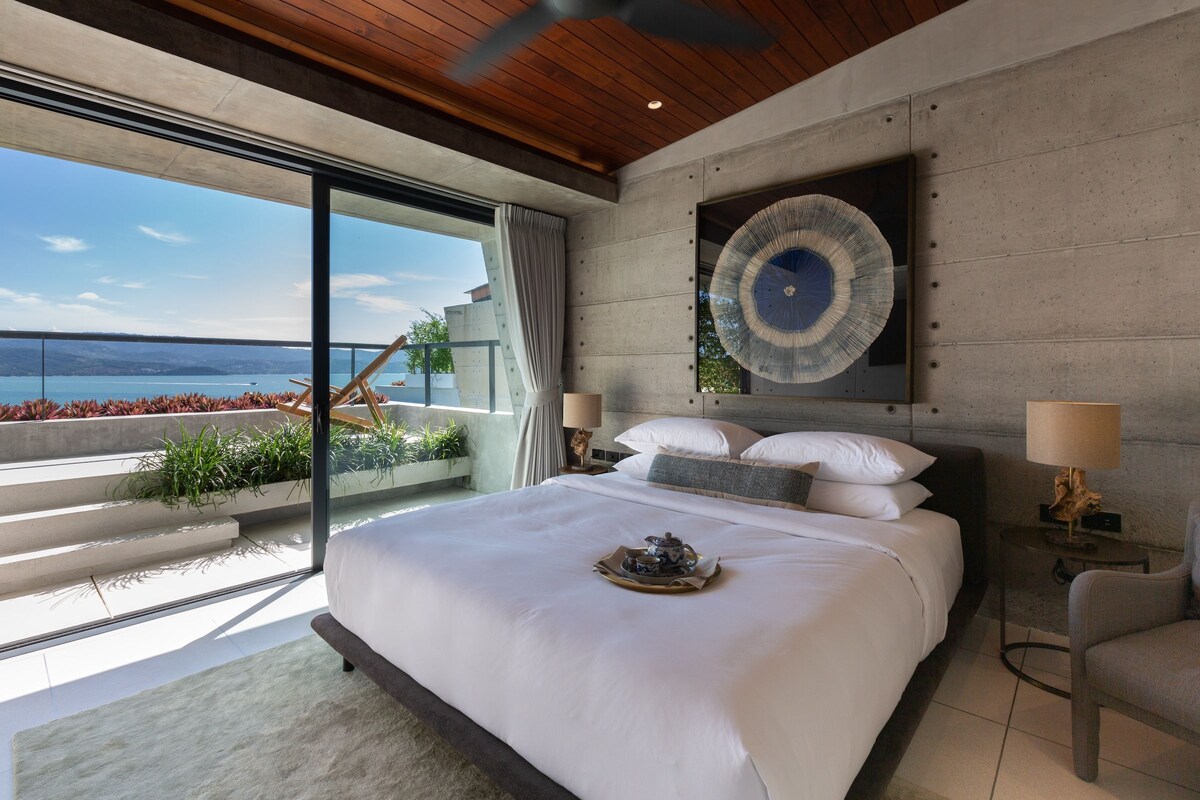A modern bedroom with a large bed, a view of the sea, and a dining table with a tray.
