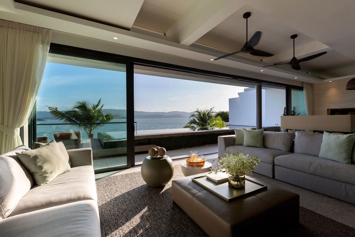 A modern living room with a large window overlooking a body of water and mountains. There are two sofas, a coffee table, and a floor lamp.
