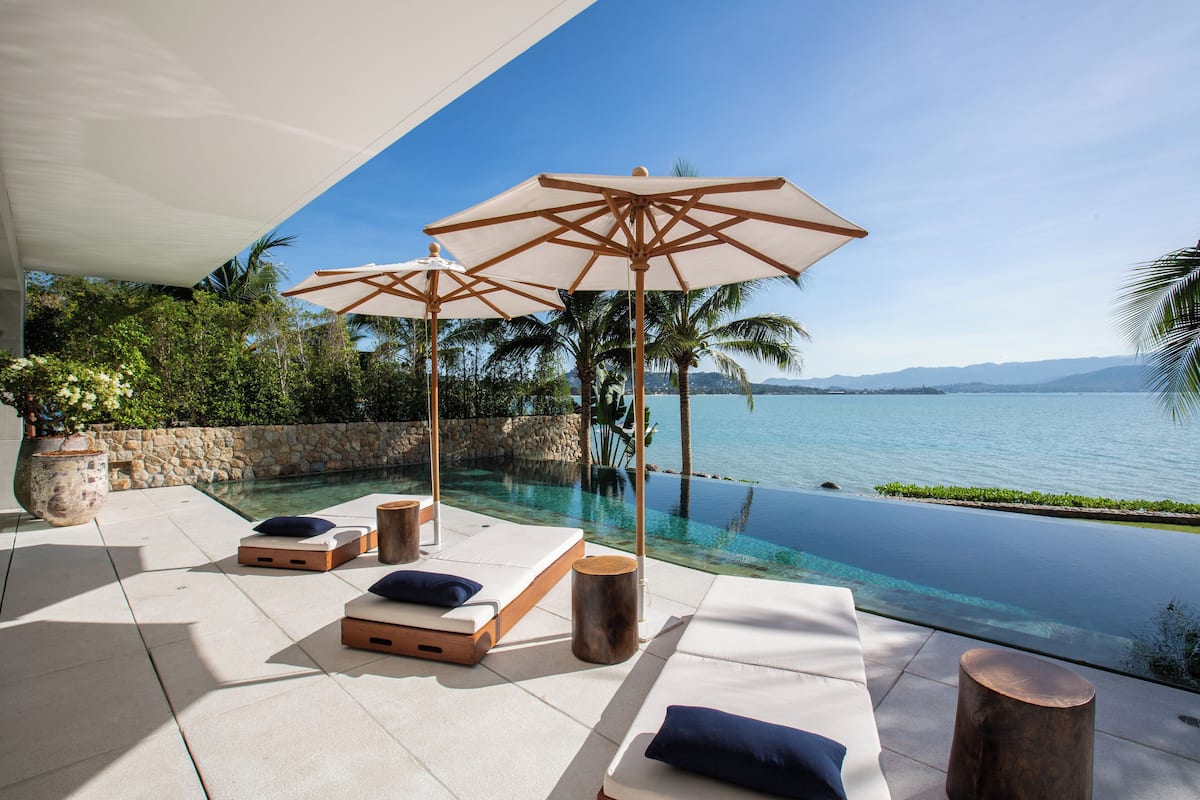 An infinity pool with lounge chairs and umbrellas, surrounded by palm trees and a view of the ocean.