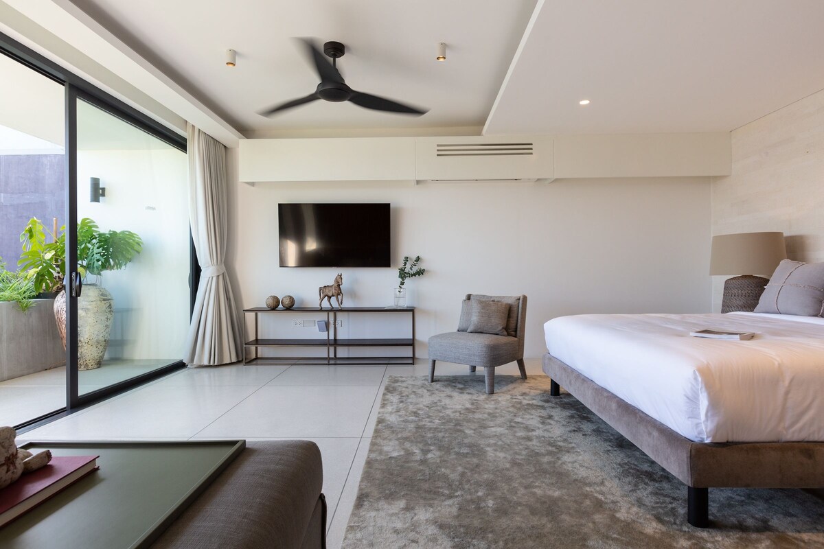 Une chambre moderne avec un grand lit, un ventilateur de plafond, une télévision et une porte-fenêtre coulissante donnant sur un balcon agrémenté de plantes.