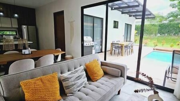 Un salon moderne avec un canapé gris, des coussins jaunes et une vue sur une piscine et un espace repas extérieur.