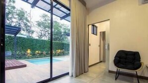 Une chambre d’hôtel moderne dotée d’une porte-fenêtre coulissante donnant sur un espace piscine, d’un fauteuil noir rembourré et d’une petite table en bois.