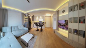Una sala moderna con un sofá, una mesa de centro y un televisor. Cuenta con una zona de cocina con taburetes de bar y una mesa de comedor. El piso es de madera y la pared blanca tiene estanterías.