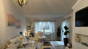 A living room with a dining area, a TV mounted on the wall, and a large window with curtains. - Stylish 2nd floor Ocean Edge condo (Brewster)