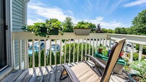 A wooden deck with a chair and potted plants, overlooking a street with parked cars and greenery. - Stylish 2nd floor Ocean Edge condo (Brewster)