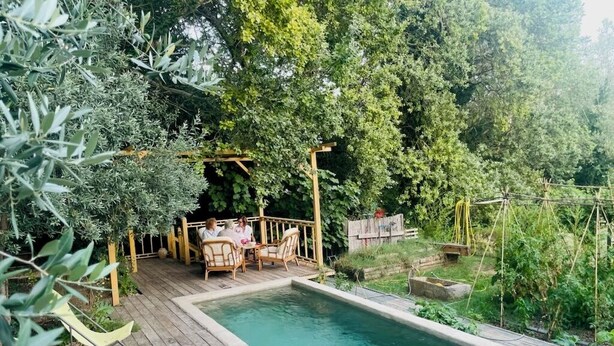 A wooden deck with a swimming pool, surrounded by lush greenery and a wooden pergola with seating. -  A quiet natural setting, a comfortable 60M2 apartment with swimming pool & bbq. (Saint etienne du gres)