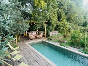 A wooden deck with a swimming pool, surrounded by lush greenery and a wooden pergola with seating. -  A quiet natural setting, a comfortable 60M2 apartment with swimming pool & bbq. (Saint etienne du gres)