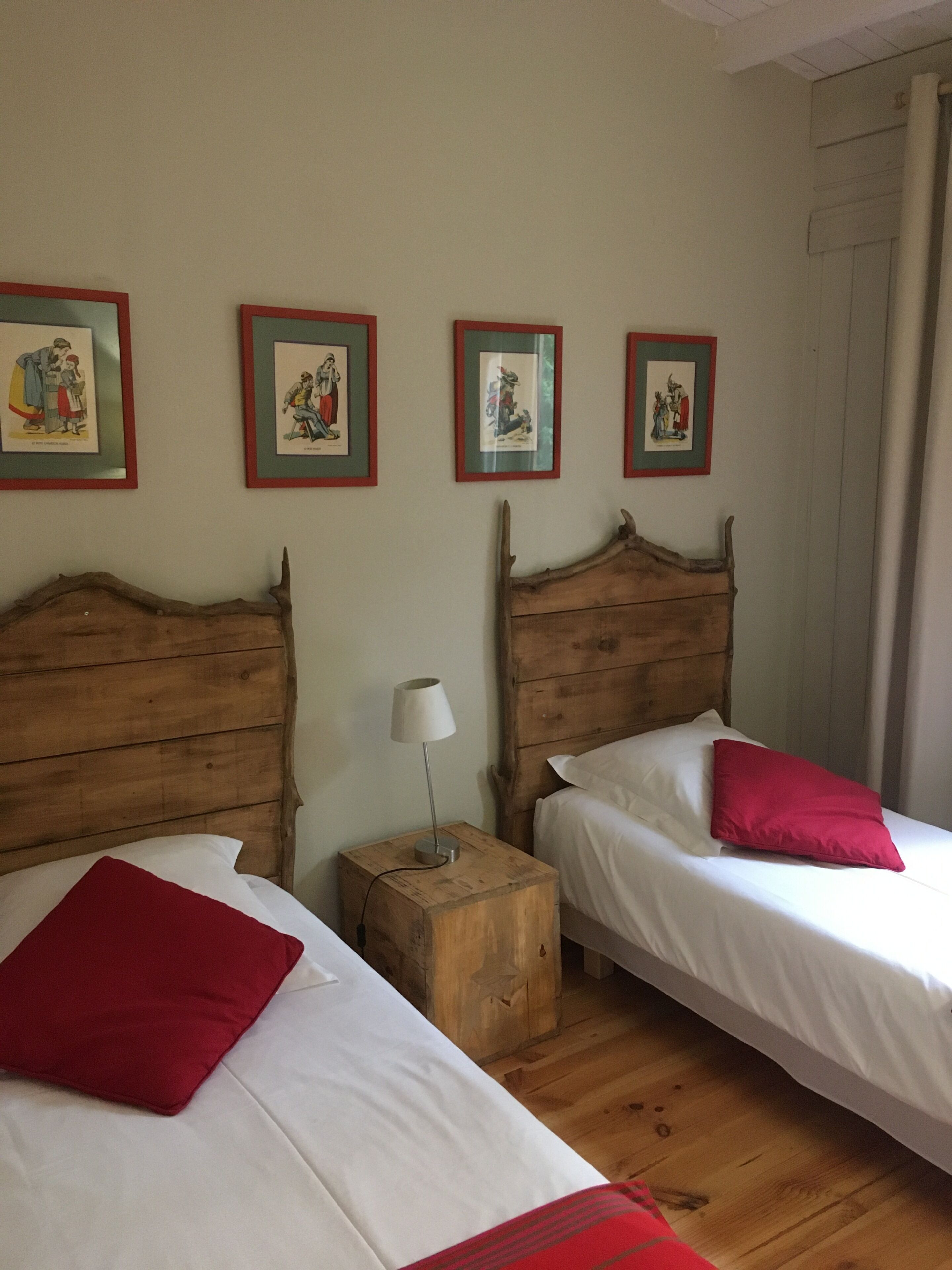 Two wooden beds with white linens and red pillows, a wooden nightstand with a lamp, and four framed pictures on the wall.