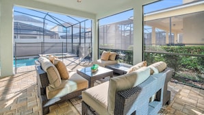 A sunlit patio with wicker furniture, a pool, and large windows offering views of the surrounding area.