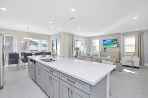 A modern kitchen with a large island, dining area with chairs, and a living room with a sofa and TV.