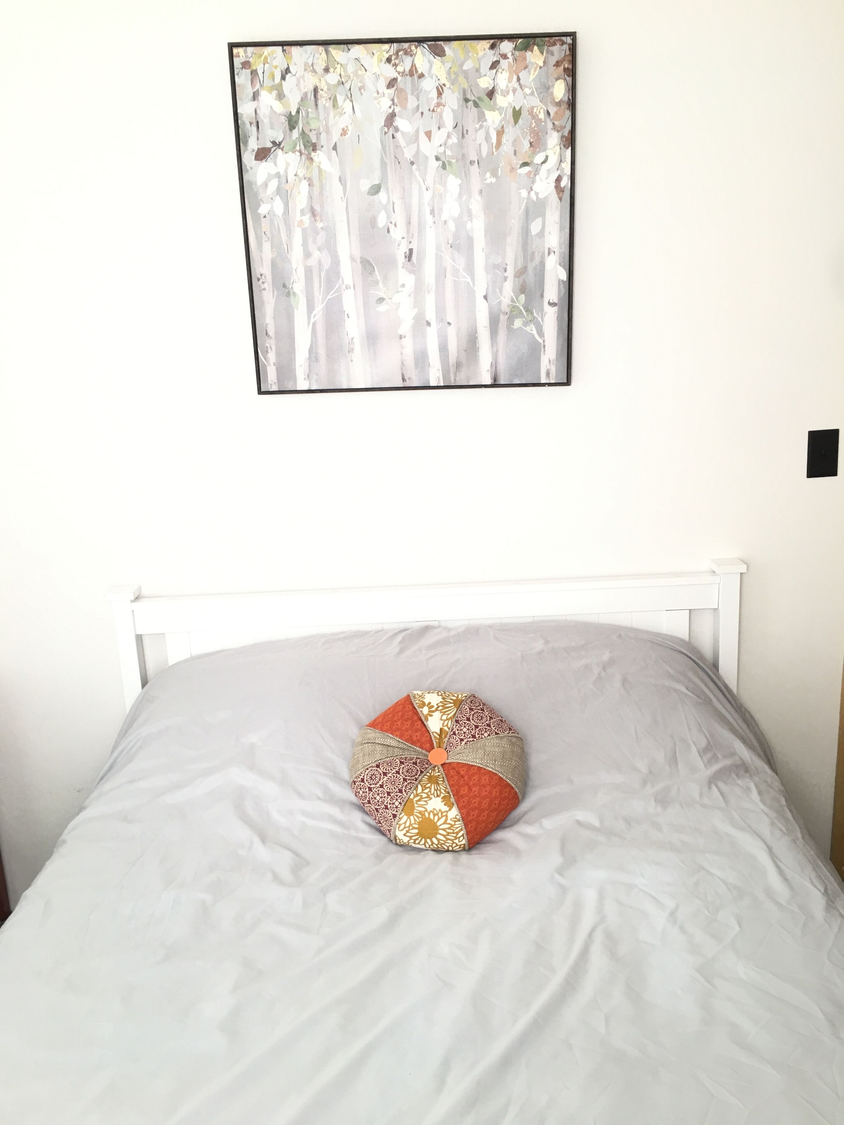 Uma cama com cabeceira branca, colcha cinza e duas almofadas decorativas. Uma obra de arte emoldurada com árvores está pendurada na parede acima da cama.