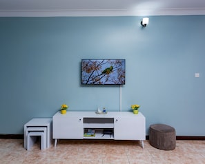 Une pièce avec un mur bleu, un meuble blanc sur lequel est posé un téléviseur à écran plat, une petite table blanche et un pouf rond.