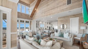 A spacious living area with a high ceiling, large windows, and a view of the ocean. - 5BR Designer Home with Gulf Views, Elevator + Beach Access | Pelican Pointe (Destin)