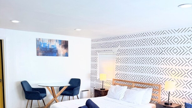A modern hotel room with a wooden bed, a desk with chairs, a patterned wall, and a window with blinds. - Sunshine Escape Guesthouse ☀ (Orlando)