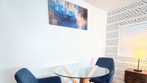 A modern dining area with a glass table and two blue chairs, a painting on the wall, and a patterned wall behind a window. - Sunshine Escape Guesthouse ☀ (Orlando)