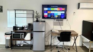 A modern office space with a flat-screen TV mounted on the wall displaying various apps and channels. There is a desk with a chair, a refrigerator, a microwave, and a trash bin. - Sunshine Escape Guesthouse ☀ (Orlando)