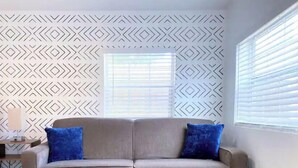 A beige sofa with two blue cushions in a room with patterned wallpaper and a window with blinds. - Sunshine Escape Guesthouse ☀ (Orlando)