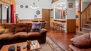 Una sala de estar acogedora con un sofá de cuero marrón, una mesa de centro de madera y un comedor con sillas de madera, además de una pequeña cocina al fondo.