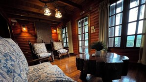 A cozy log cabin interior with a patterned sofa, two armchairs, a round wooden table, and large windows with curtains.