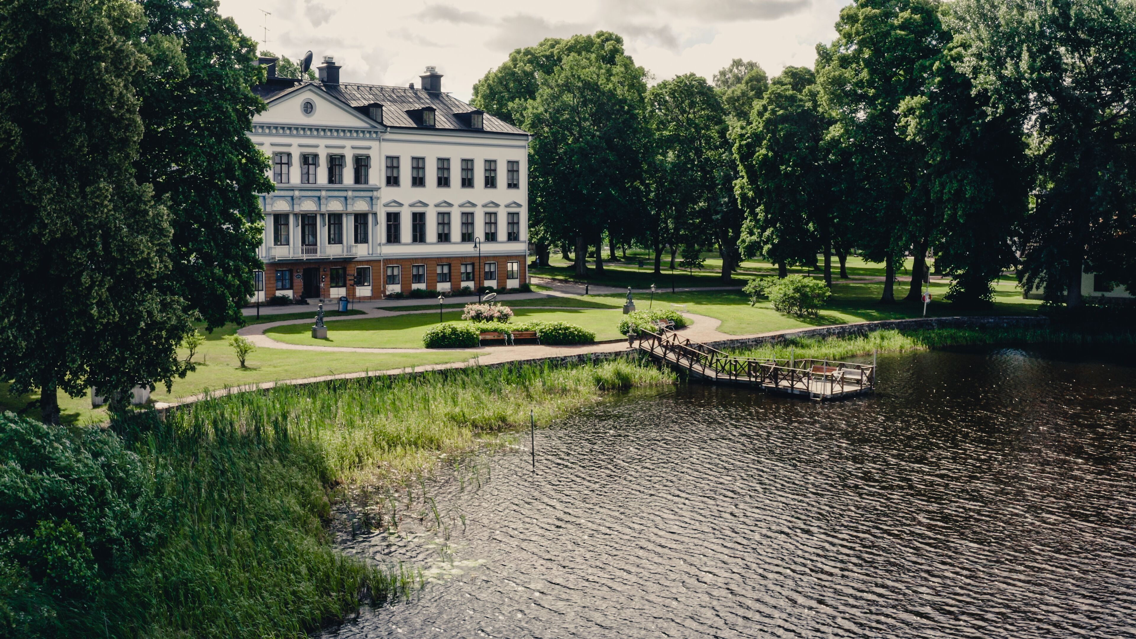 En stor, historisk byggnad med en symmetrisk fasad, en välskött gräsmatta och en bro över ett stilla vattenområde.