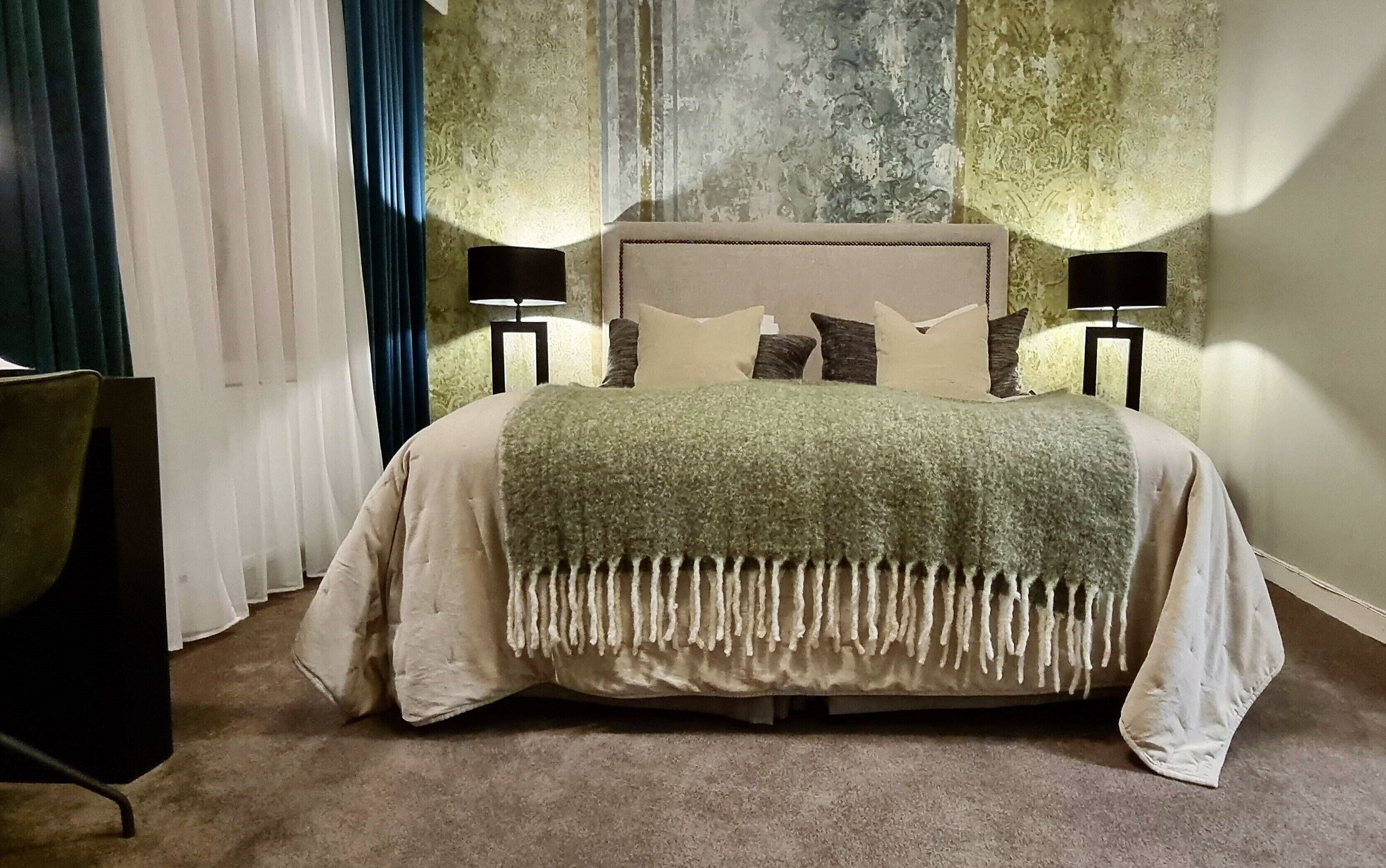 A neatly made bed with a green blanket and pillows, a bedside table with a lamp, and a chair.