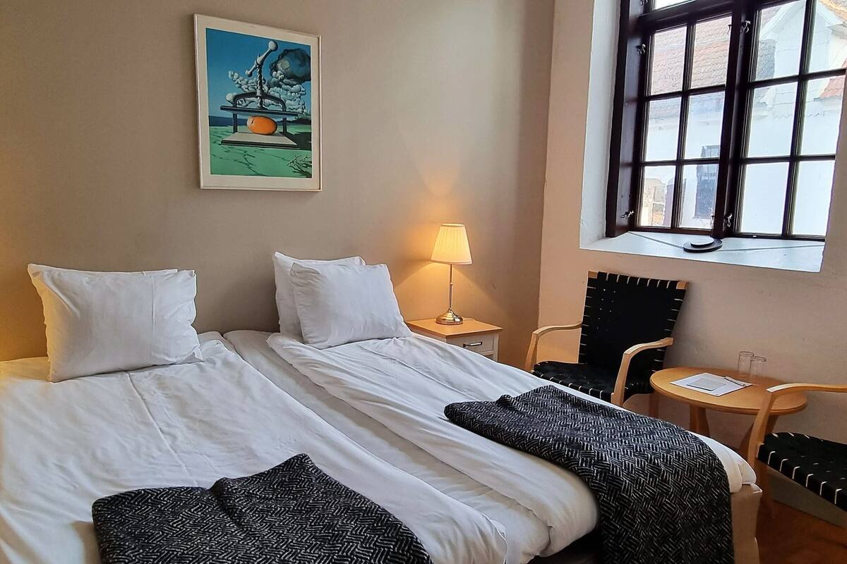 Quarto de hotel com uma cama de casal, duas mesas de cabeceira com luminárias, uma mesinha redonda, uma cadeira e uma janela com persianas.