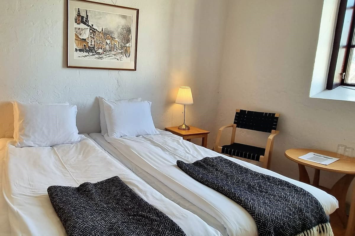 Quarto de hotel com cama, duas luminárias de cabeceira, uma cadeira, uma mesinha e um quadro na parede.