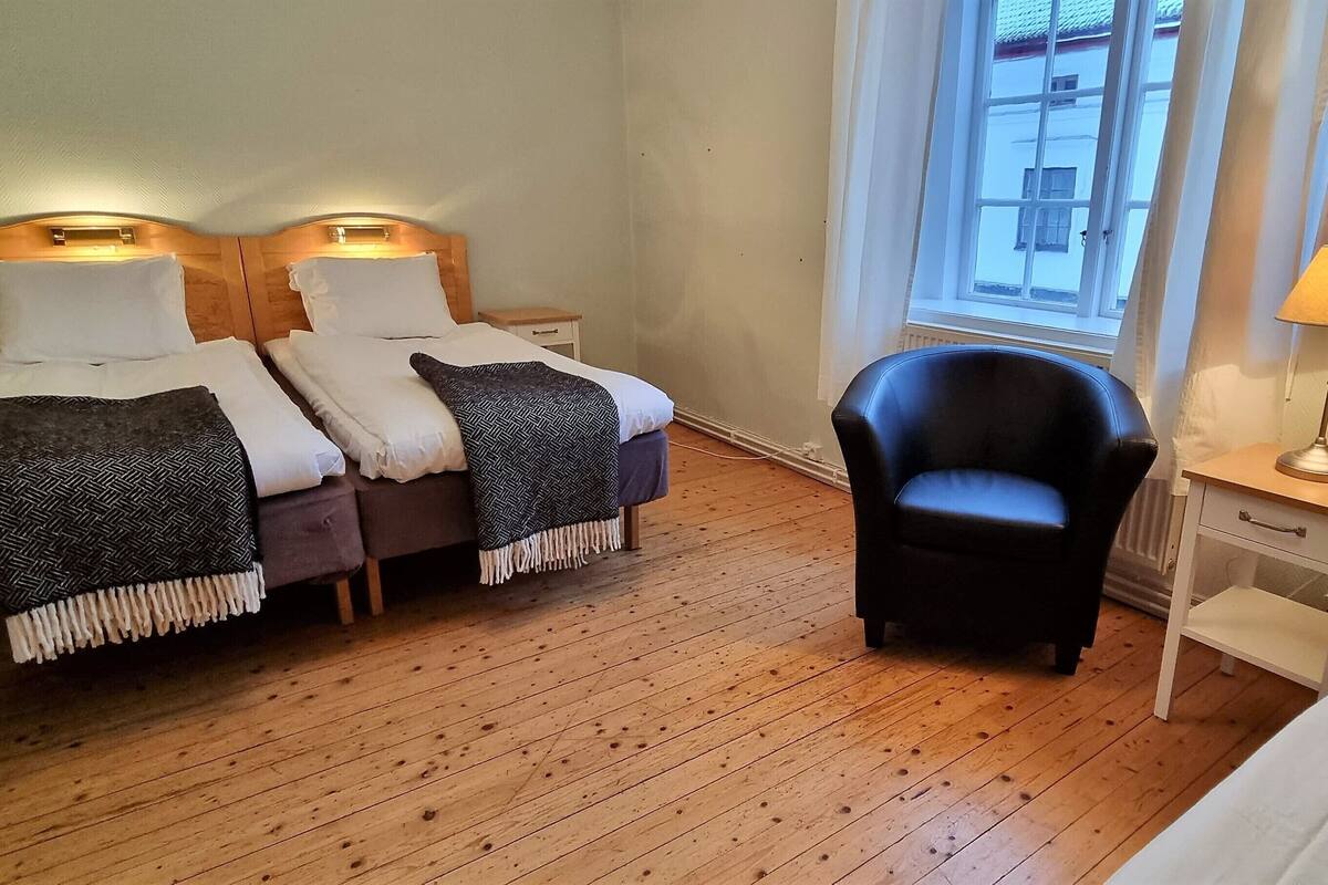 Quarto com duas camas, uma cadeira, uma mesa de cabeceira, abajur e janela com cortinas.