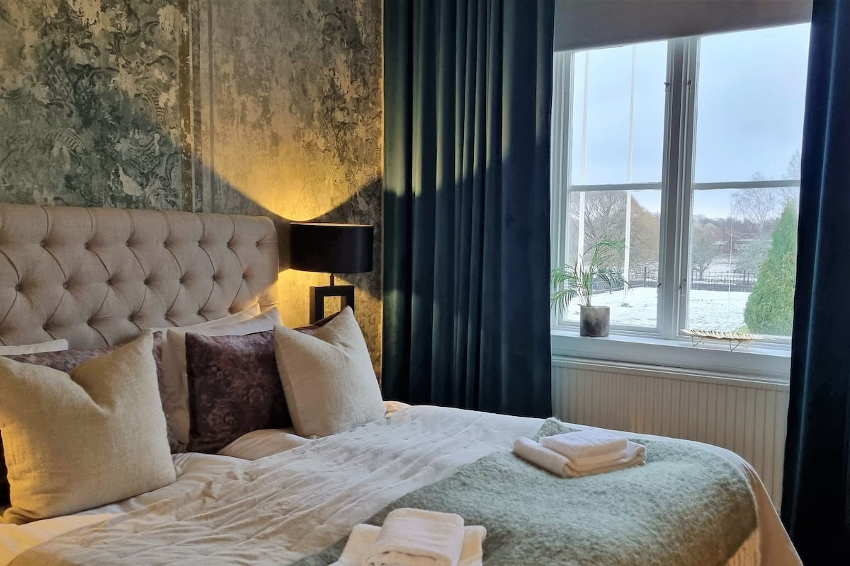 Uma cama bem arrumada com almofadas e um cobertor, uma luminária em uma mesa de cabeceira e vista para um jardim nevado pela janela.
