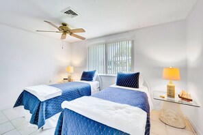 Two single beds with bedside tables and lamps, ceiling fan, and a large window with vertical blinds.