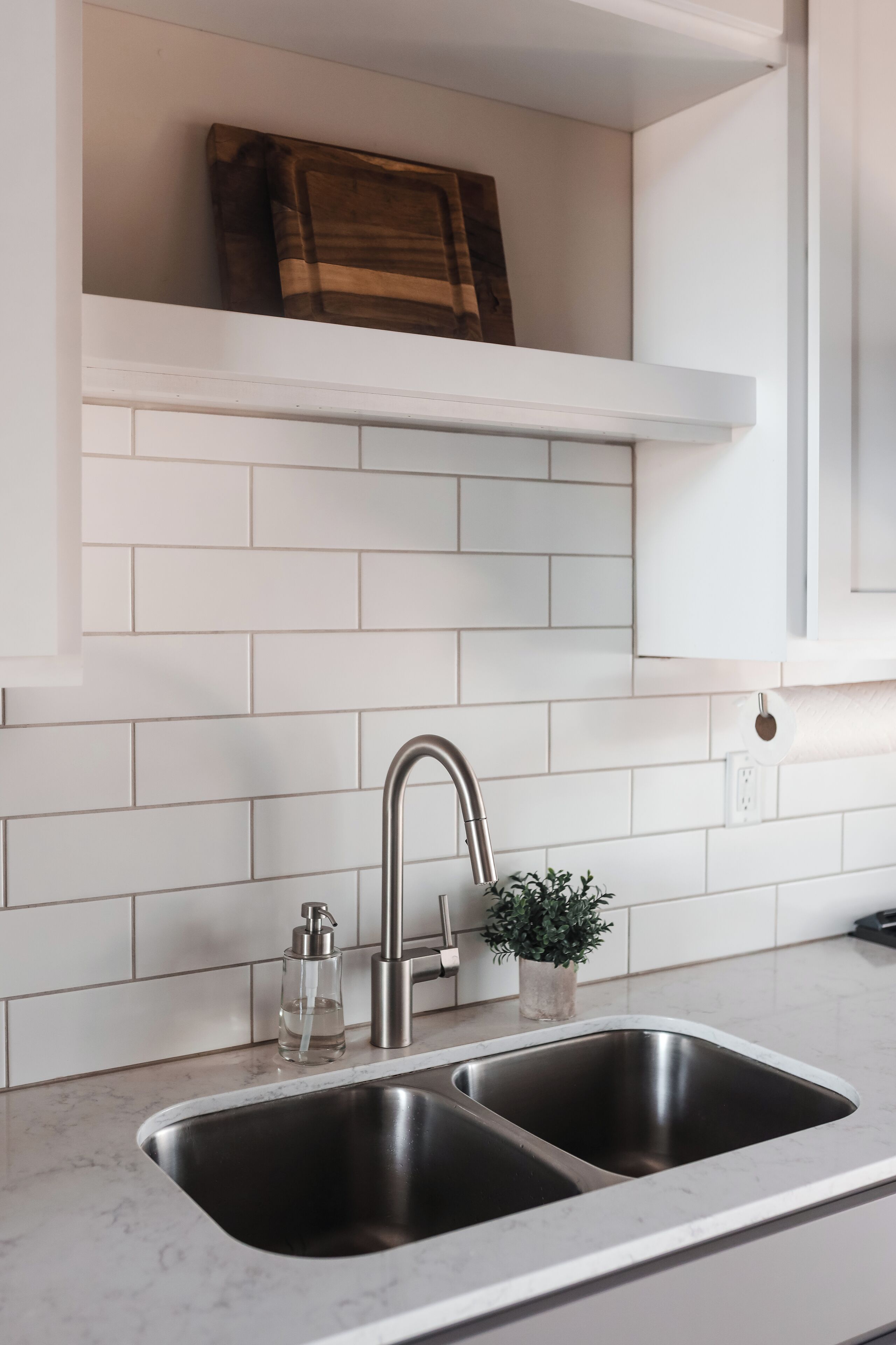 A kitchen with white subway tiles, a stainless steel sink, a faucet, a soap dispenser, and a small potted plant.