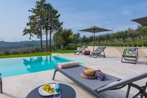 Uma área à beira da piscina com espreguiçadeiras, uma mesa com uma garrafa e uvas, e vista para o campo.