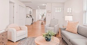 A modern living room with a sofa, armchair, and coffee table, a kitchen in the background, and a hallway leading to a dining area.