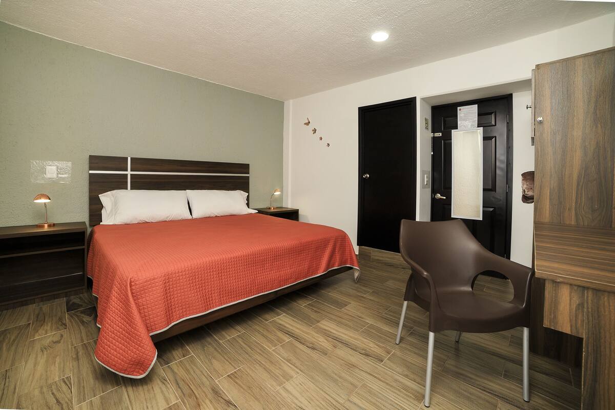 A hotel room with a bed, a chair, a nightstand, and a door.