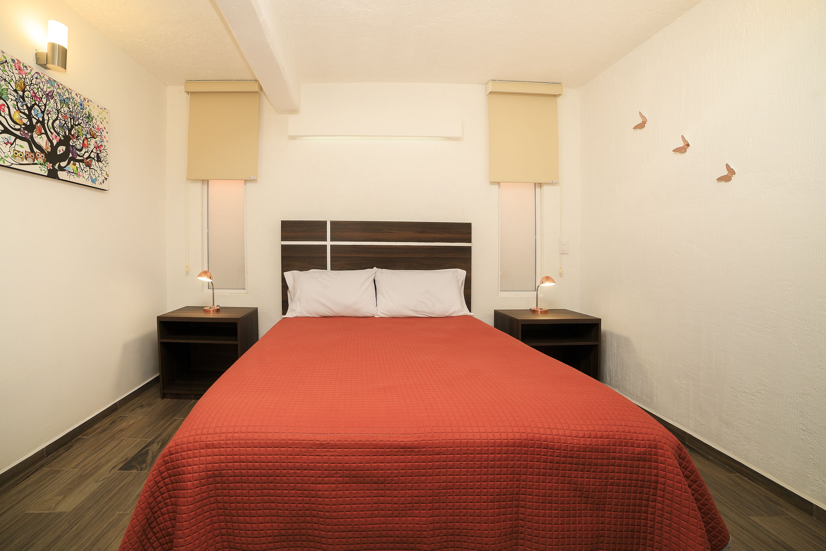 Una cama bien hecha con una colcha roja, dos mesitas de noche con lámparas, una obra de arte en la pared y una lámpara del techo.
