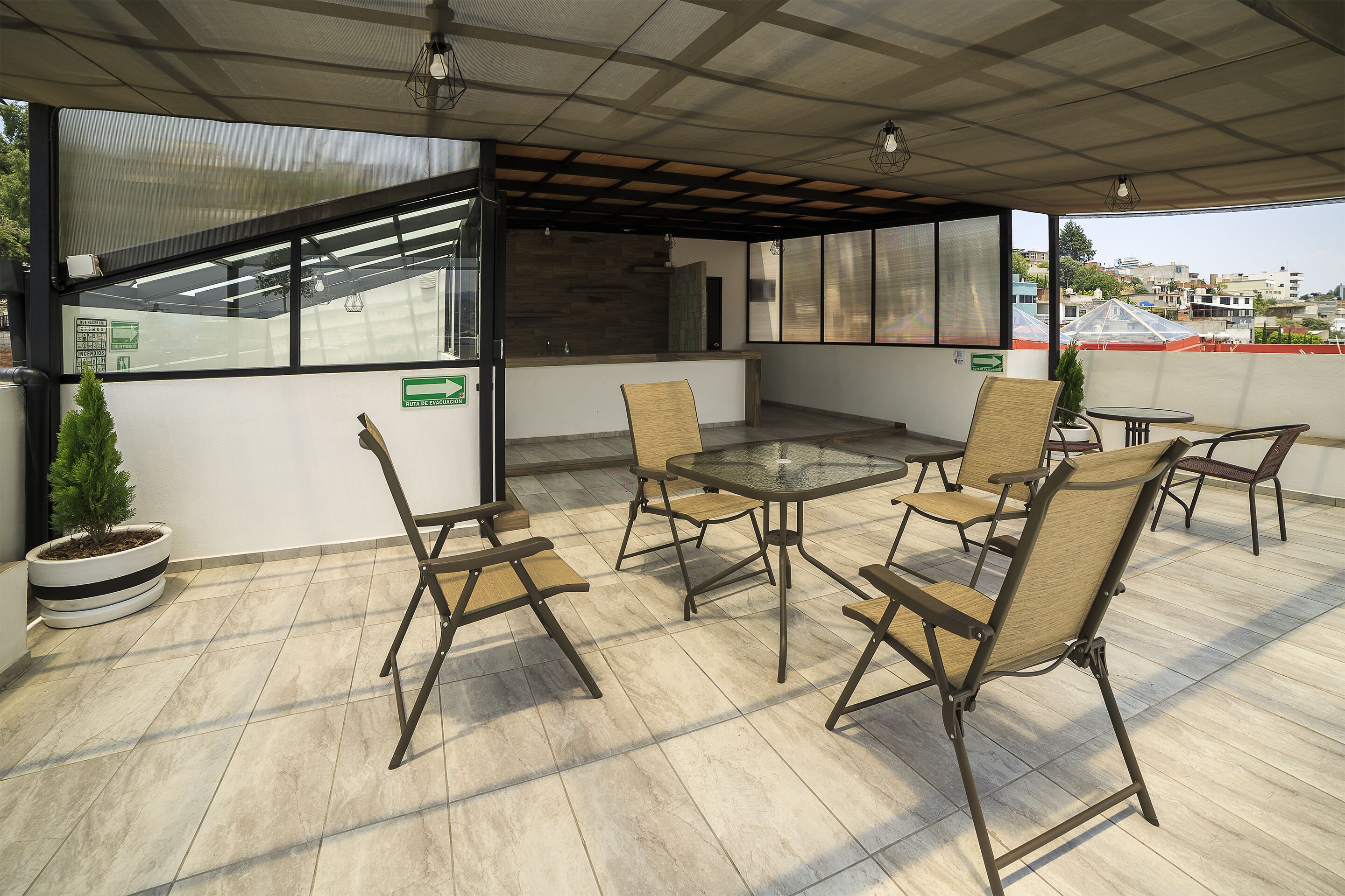 A covered outdoor seating area with a glass table and chairs, a small potted plant, and a view of buildings in the background.