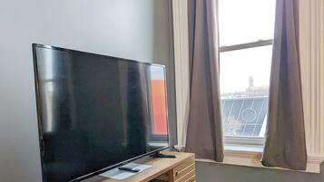 A flat-screen TV on a wooden stand in a room with a large window and curtains.