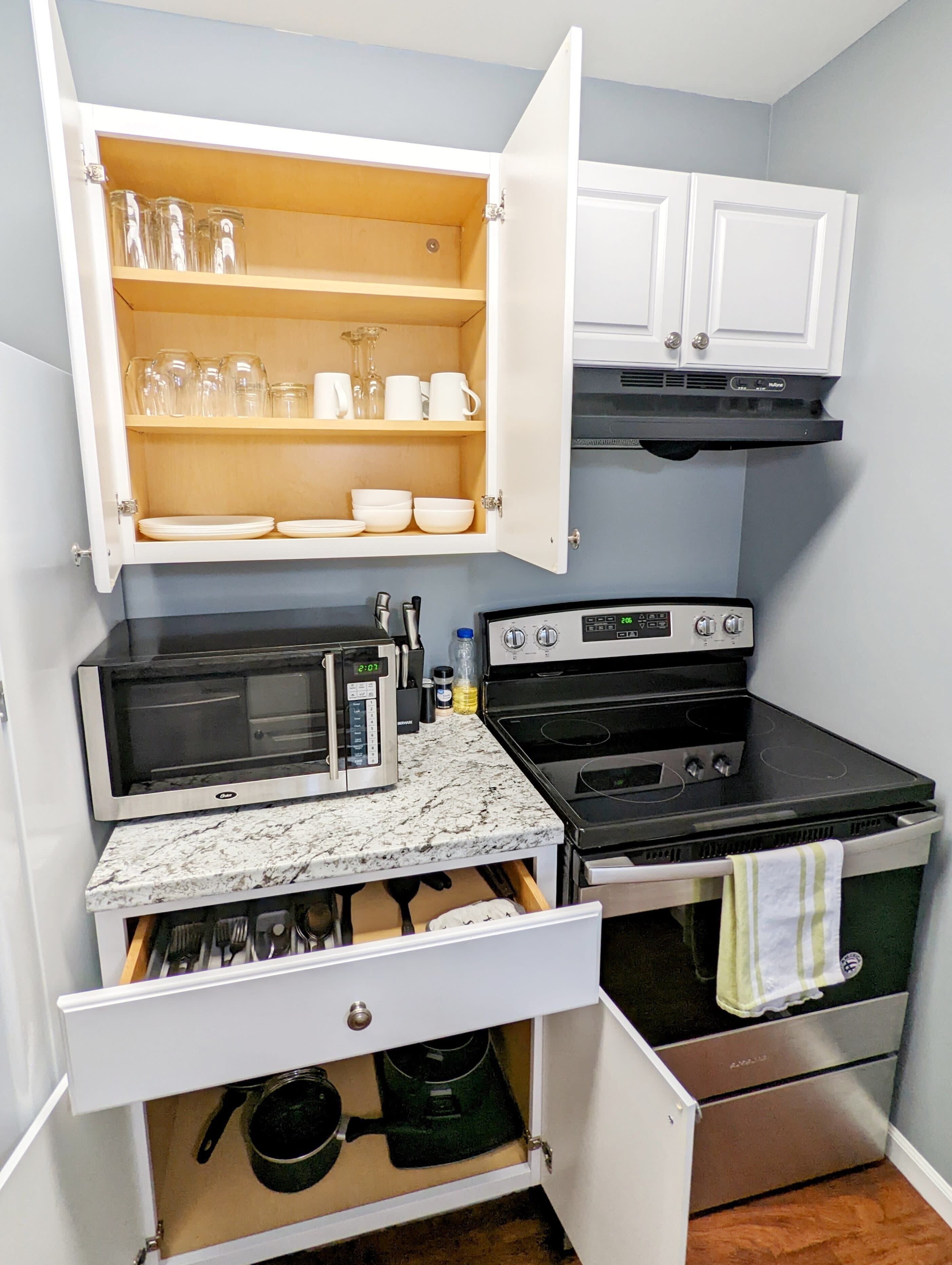 A compact kitchen with a microwave, stove, and oven, a cabinet with glassware, and a countertop with utensils.
