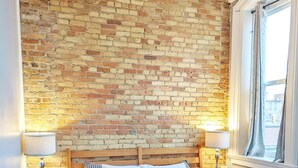 Un dormitorio con pared de ladrillo, cabecera de madera, dos mesitas de noche con lámparas, cama con ropa de cama a rayas y una ventana con cortinas.