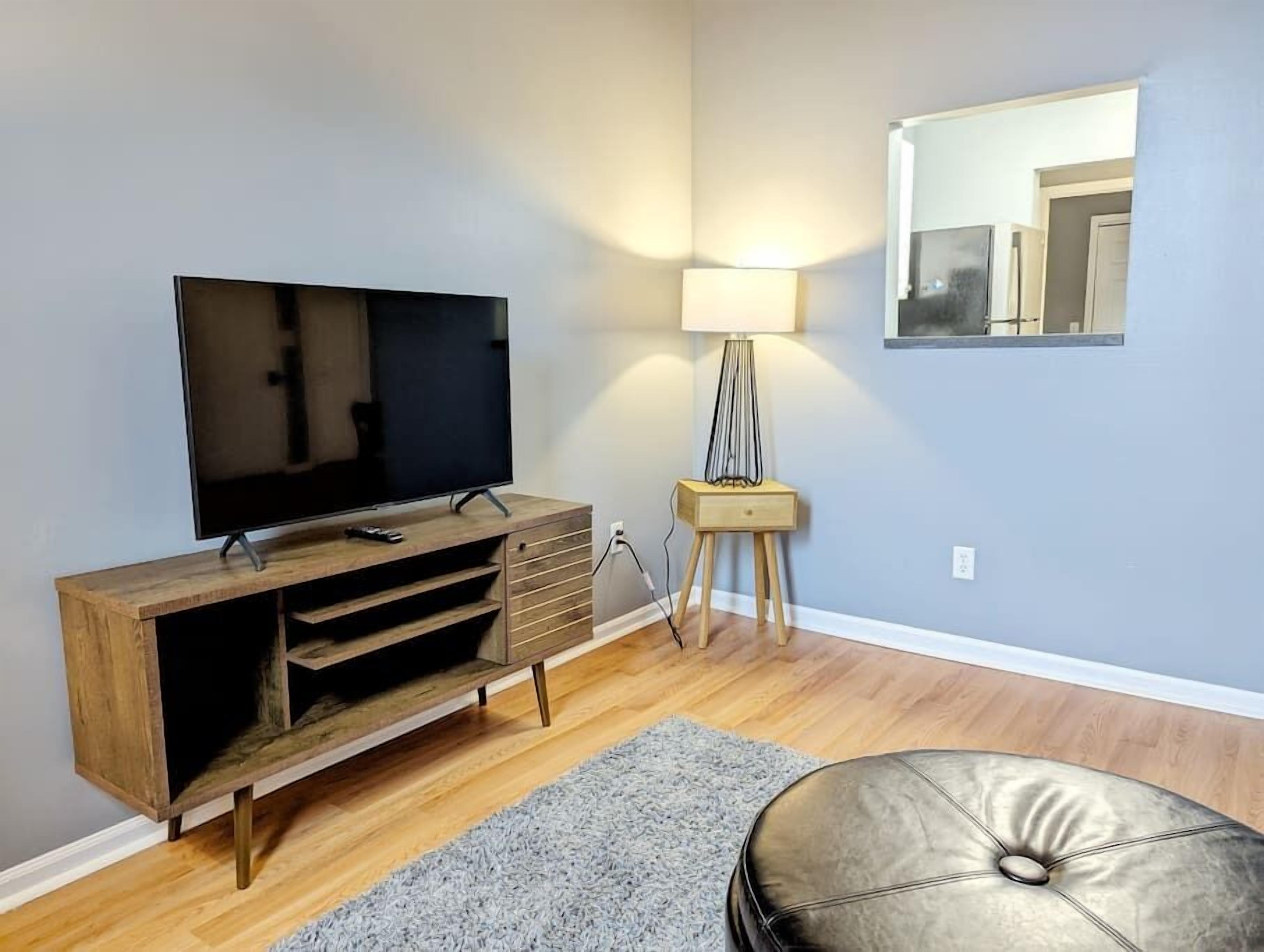 A living room with a flat-screen TV on a wooden stand, a small side table with a lamp, a grey rug, and a leather ottoman.