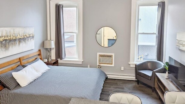 A hotel room with a bed, a desk, a chair, a mirror, and two windows with curtains. - Tanner Bldg Apt 5 - Studio (Downtown) (Bay City)