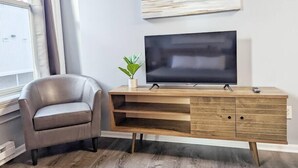 A modern living room with a leather armchair, a wooden entertainment unit with a flat-screen TV, and a potted plant. - Tanner Bldg Apt 5 - Studio (Downtown) (Bay City)
