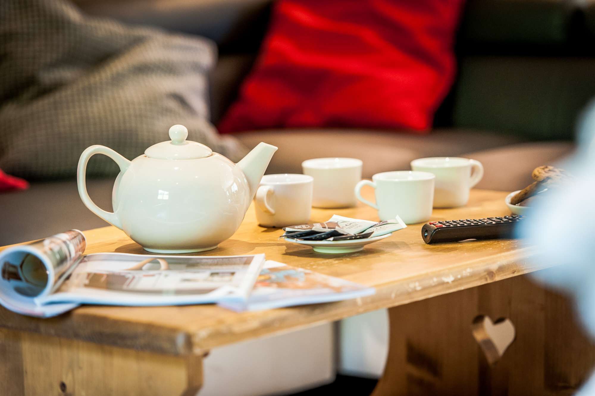 Una mesa de madera con un hervidor blanco, varias tazas blancas, un control remoto y algunos saquetes de té.