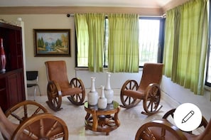 Relax in a traditional room with a wooden rocking chair, matching chair, and a small table, surrounded by a painting and green curtains.