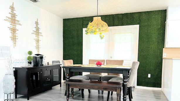 A dining area with a wooden table, chairs, and a bench. There is a black cabinet with a coffee machine and a sign that says "COFFEE". The walls are white with a green accent wall.