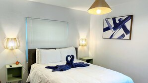 A neatly made bed with white bedding, two bedside lamps, a wall-mounted abstract painting, and a window with blinds.
