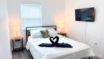 A hotel room with a bed, two bedside lamps, a TV mounted on the wall, and a window with blinds.
