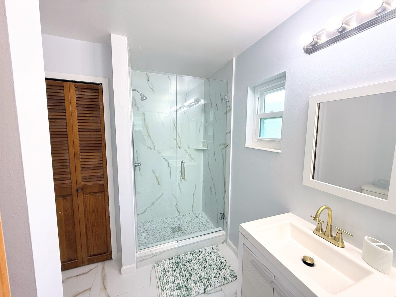 Un baño con ducha envuelta en vidrio, un lavabo blanco con grifo dorado y una puerta de madera.